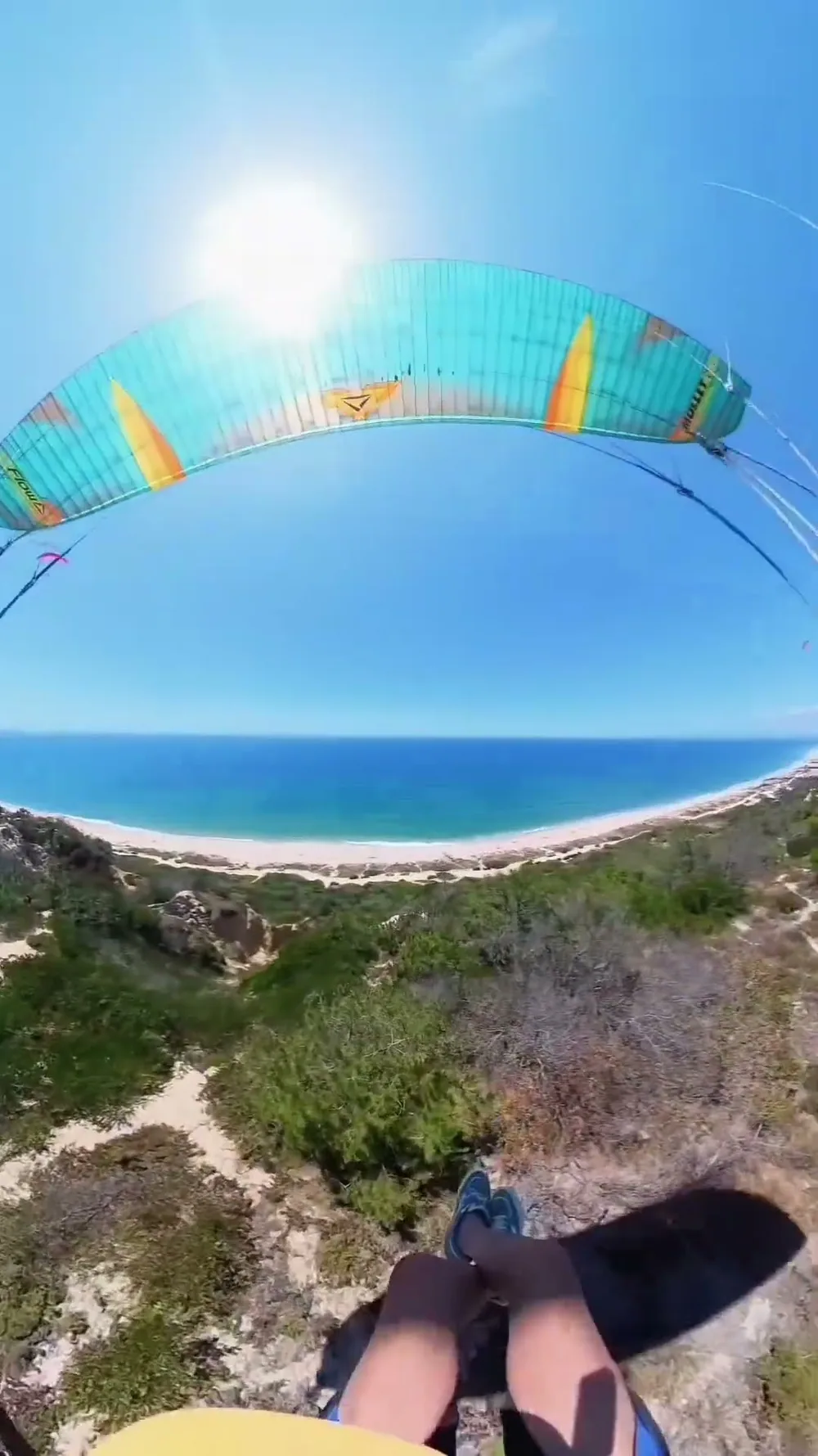 La vista del piloto bajo un parakite Flow turquesa, volando bajo sobre la vegetación dunar de la Fonte da Telha, con la playa y el Atlántico al fondo.