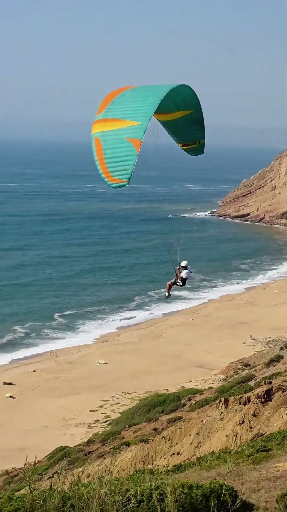 Un parakite turquesa y naranja volando bajo sobre el arenal de la Praia da Gralha, con el Atlántico a la izquierda y los acantilados ocres a la derecha.