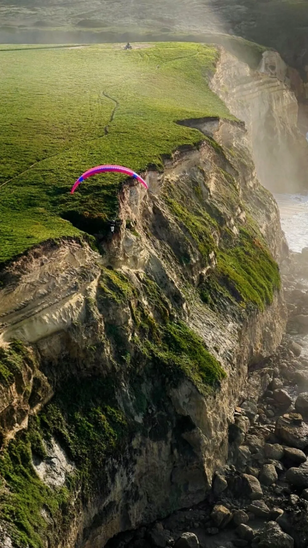 Parapente rosa planeando junto al acantilado verde de la Praia das Bicas, con bruma marina y rocas abajo.