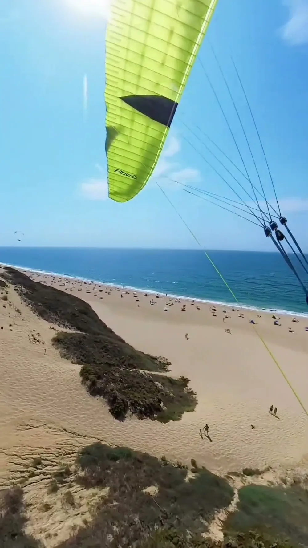 La vista del piloto bajo un ala Flow verde flúor, volando junto al acantilado de la Praia do Meco sobre un arenal lleno de bañistas, con otra ala a lo lejos.